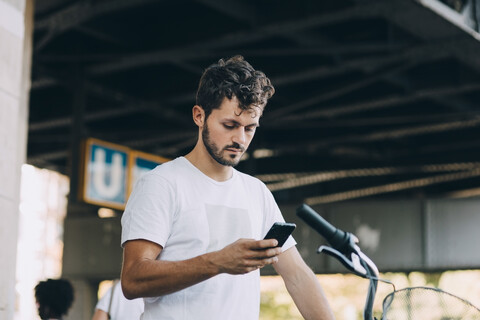 Junger Mann benutzt sein Smartphone, während er mit dem Fahrrad unter einer Brücke in der Stadt steht, lizenzfreies Stockfoto