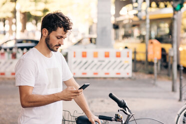 Junger Mann, der ein Mobiltelefon benutzt, während er mit dem Fahrrad auf einer Straße in der Stadt steht - MASF10356