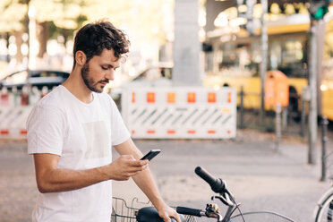 Young man using mobile phone while standing by bicycle on street in city - MASF10356