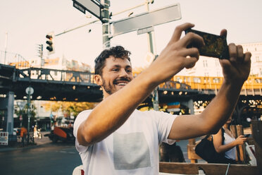 Smiling young man photographing through smart phone while standing on street in city - MASF10363