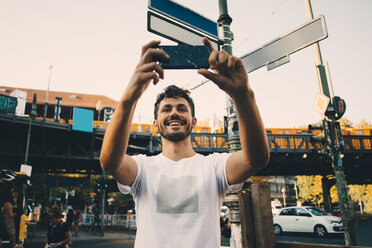 Lächelnder junger Mann, der mit seinem Mobiltelefon fotografiert, während er auf der Straße in der Stadt steht - MASF10365