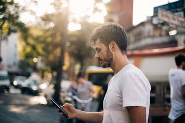 Seitenansicht eines jungen Mannes, der ein Mobiltelefon benutzt, während er auf einer Straße in der Stadt steht - MASF10366