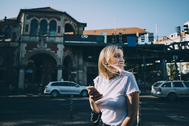 Junge Frau schaut über die Schulter, während sie auf der Straße in der Stadt ein Mobiltelefon hält - MASF10383