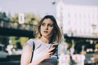 Thoughtful young woman holding mobile phone while standing on street in city - MASF10384