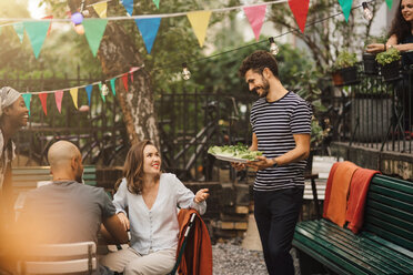 Lächelnde junge Frau im Gespräch mit einem männlichen Freund, der Essen bei einer Gartenparty trägt - MASF10406