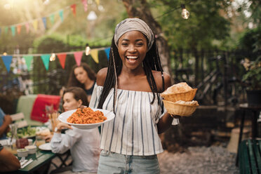 Portrait of smiling young woman carrying food while standing in backyard during garden party - MASF10413