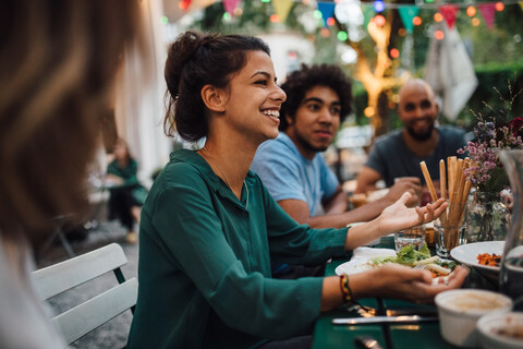 Lächelnde junge Frauen gestikulieren beim Sitzen mit Freunden am Tisch während einer Dinnerparty, lizenzfreies Stockfoto