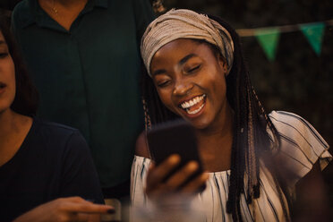 Junge Frau lacht, während sie auf ihr Handy schaut, während einer Dinnerparty mit Freunden im Hinterhof - MASF10444