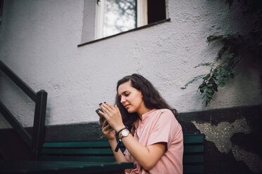 Young woman using mobile phone while sitting on bench outside house - MASF10485