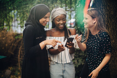 Lächelnde multiethnische Freundinnen schauen auf ihr Handy, während sie im Hinterhof stehen - MASF10504