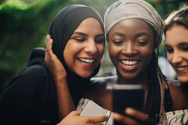 Lächelnde multiethnische Freunde, die auf ihr Handy schauen, während sie im Hinterhof stehen - MASF10509