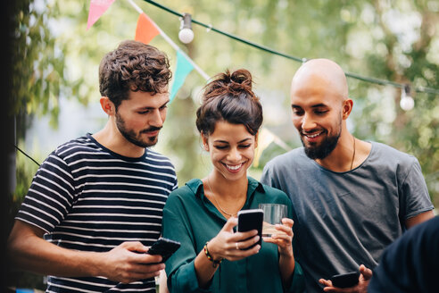 Glückliche junge Frau zeigt männlichen Freunden ihr Handy, während sie auf dem Balkon einer Party steht - MASF10516