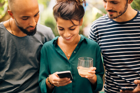 Lächelnde junge Frau zeigt männlichen Freunden ihr Handy, während sie im Freien steht - MASF10538