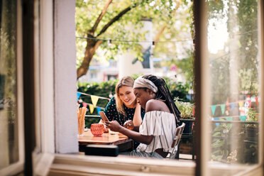 Multi-ethnic female friends looking at mobile phone seen through window - MASF10541