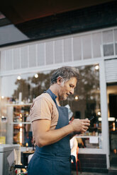 Seitenansicht eines männlichen Besitzers, der vor einem Feinkostladen ein Mobiltelefon benutzt - MASF10567