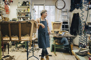 Älterer männlicher Handwerker, der neben einem unvollständigen Sofa in einer Polsterwerkstatt steht und wegschaut - MASF10601