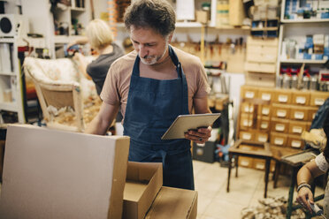 Männlicher Handwerker, der ein digitales Tablet hält, während er einen Karton in einer Werkstatt öffnet - MASF10618