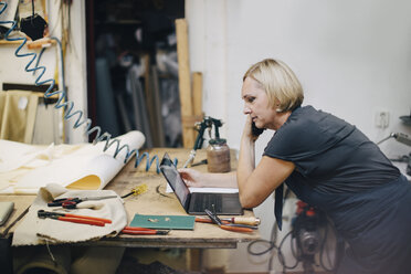 Seitenansicht einer reifen Polstermöbelherstellerin, die auf einen Laptop schaut, während sie in einer Werkstatt telefoniert - MASF10625