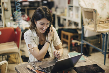 Lächelnde Polstermöbelherstellerin, die einen Laptop benutzt, während sie an einer Werkbank in einer Werkstatt telefoniert - MASF10629