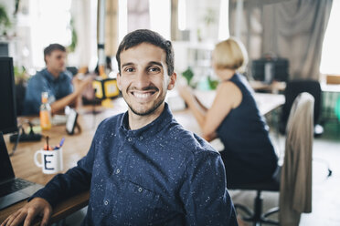 Portrait of smiling young businessman sitting at desk in creative office - MASF10653