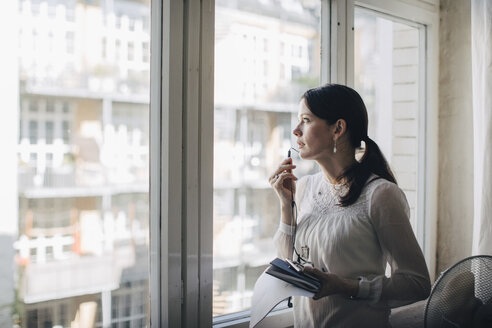 Kreative Geschäftsfrau, die über Kopfhörer spricht, während sie am Fenster im Büro steht - MASF10662