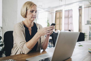 Reife Geschäftsfrau, die ein Mobiltelefon am Schreibtisch in einem kreativen Büro benutzt - MASF10669