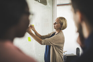 Entrepreneurs looking at female colleague sticking adhesive note on glass in creative office - MASF10703