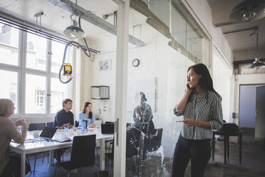 Businesswoman looking at colleagues planning strategy in board room while talking on mobile phone - MASF10710