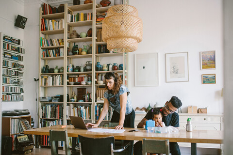 Woman using laptop while young man playing with daughter at table in house stock photo