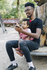 Ein kleines Mädchen benutzt ein Mobiltelefon, während es mit seinem Vater auf einem Spielplatz sitzt - MASF10779