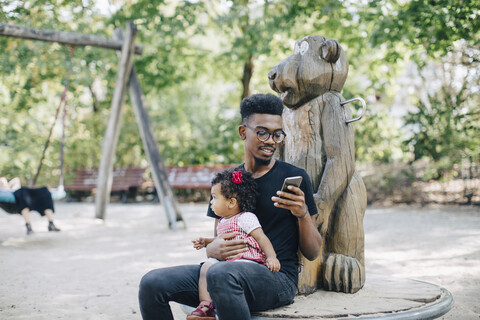 Vater benutzt Mobiltelefon, während er mit seiner Tochter auf einem Spielgerät im Freien sitzt, lizenzfreies Stockfoto