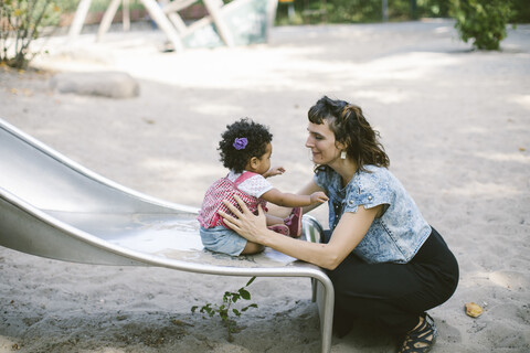 Side view of mother supporting daughter sitting on slide at playground stock photo
