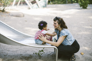Side view of mother supporting daughter sitting on slide at playground - MASF10783