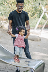 Junger Mann hält die Hände seiner Tochter auf der Rutsche eines Spielplatzes - MASF10784