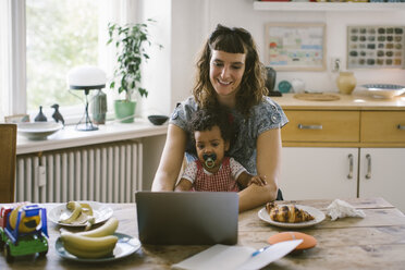 Smiling woman using laptop while sitting with daughter at dining table in house - MASF10800