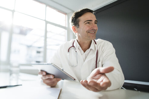 Smiling doctor sitting at desk in medical practice with tablet - JOSF02791