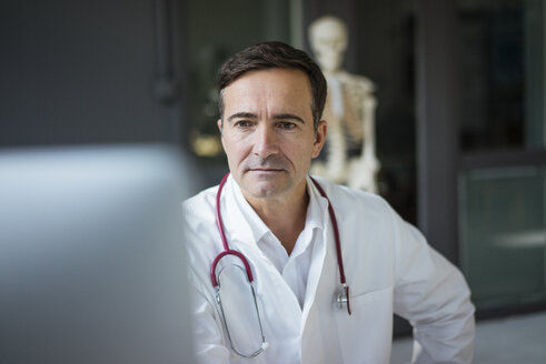Doctor in medical practice looking at computer with skeleton in background - JOSF02831