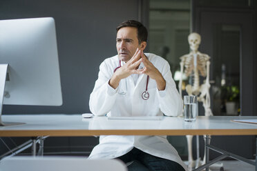 Serious doctor sitting at desk in medical practice with skeleton in background - JOSF02834