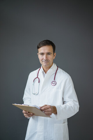 Porträt eines selbstbewussten Arztes, der ein Klemmbrett vor einer grauen Wand hält, lizenzfreies Stockfoto