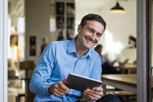 Lächelnder Geschäftsmann mit Tablet, der an einer offenen französischen Tür sitzt - JOSF02852