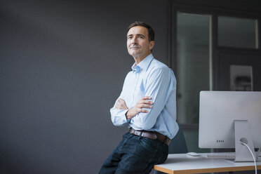 Confident businessman leaning against desk in the office - JOSF02865