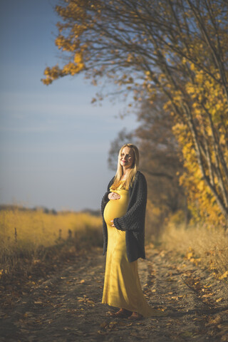 Lächelnde schwangere Frau, die auf einem Waldweg neben einem Spargelfeld im Herbst steht, lizenzfreies Stockfoto