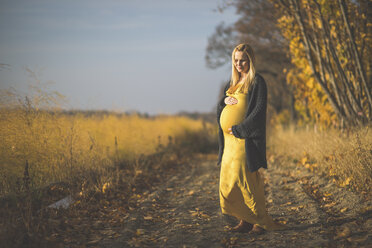Lächelnde schwangere Frau, die auf einem Waldweg neben einem Spargelfeld im Herbst steht - ASCF00938