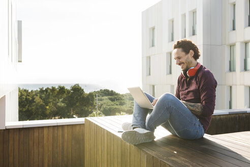 Mann sitzt im Schneidersitz auf einer Dachterrasse, benutzt einen Laptop, trägt Kopfhörer - VABF02211