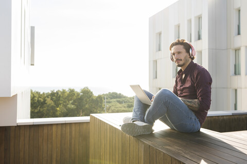 Mann sitzt im Schneidersitz auf einer Dachterrasse, benutzt einen Laptop, trägt Kopfhörer, lizenzfreies Stockfoto