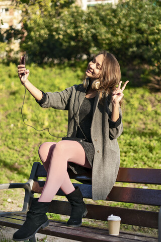 Junge Frau auf einer Parkbank sitzend, Smartphone-Selfies machend, Siegeszeichen machend, lizenzfreies Stockfoto