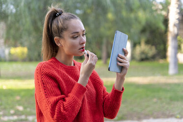 Young woman applying lipstick in apark, using digital tablet as a mirror - ERRF00645