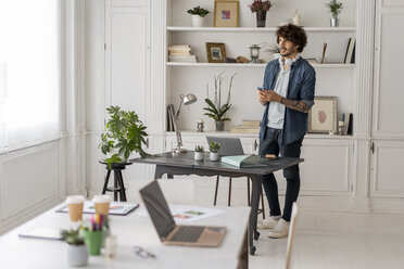 Mann arbeitet im Stehen in einem Coworking Space, benutzt ein Smartphone, hat Kopfhörer um den Hals - AFVF02224