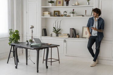 Man working standing in coworking space, using smartphone, with headphones around his neck - AFVF02225