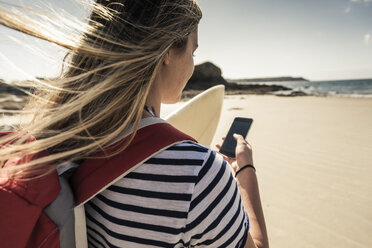 Junge Frau am Strand, mit Surfbrett, mit Smartphone - UUF16432
